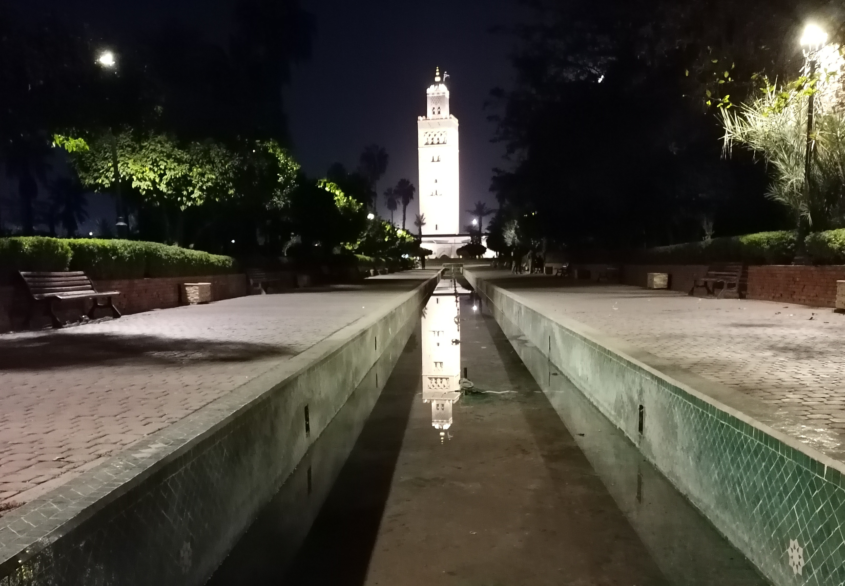 Among the trees, in the water of a canal tapering perspectivally toward the mosque in the distance, the floodlit minaret and its reflection shimmer white in the night