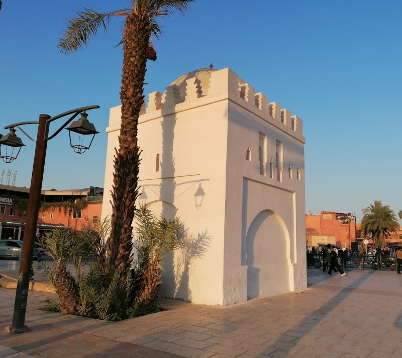 A low white cube-shaped building with a cupola on top and a stylized portal behind a palm tree.